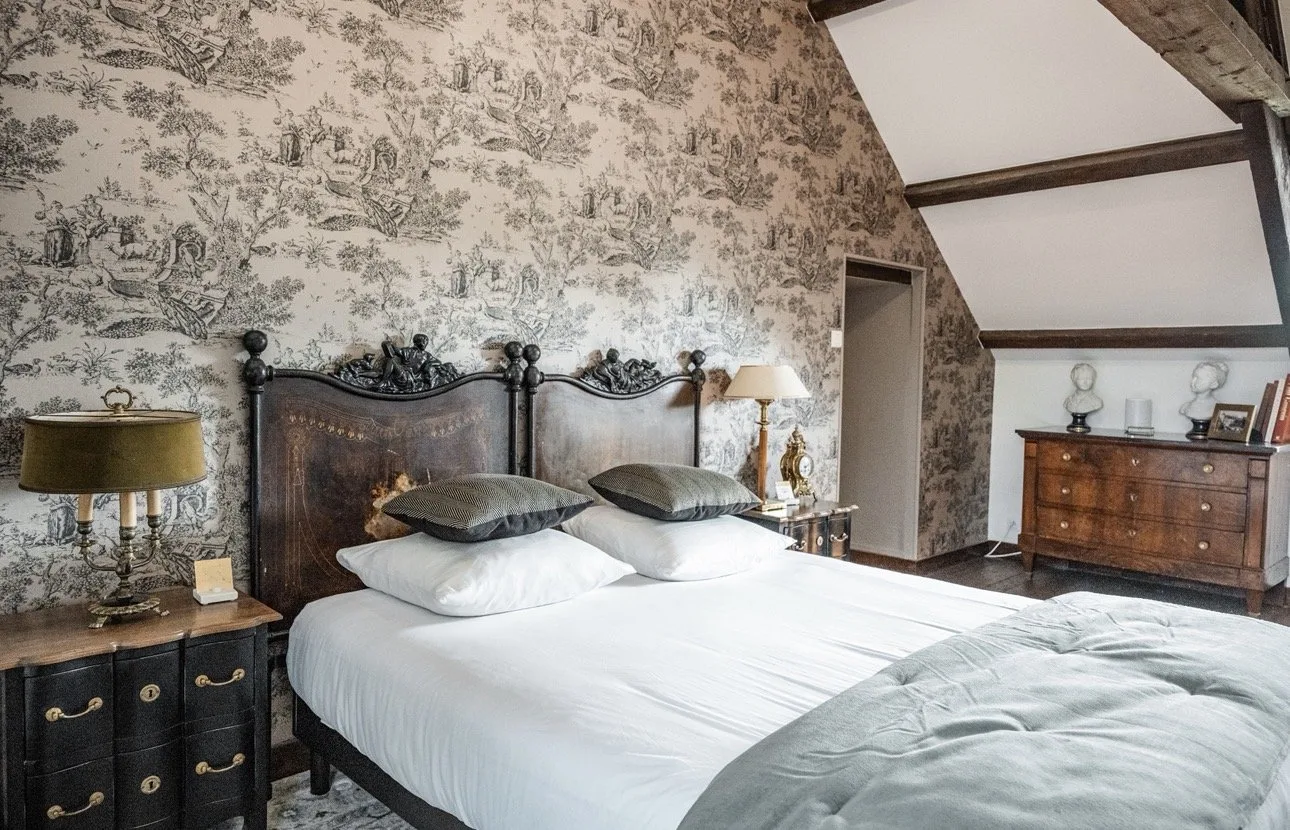 Suite La Marquise : deux têtes de lit anciennes en bois sombre sculpté devant un papier peint toile de Jouy noir et blanc, avec chevets noirs et pendule dorée.