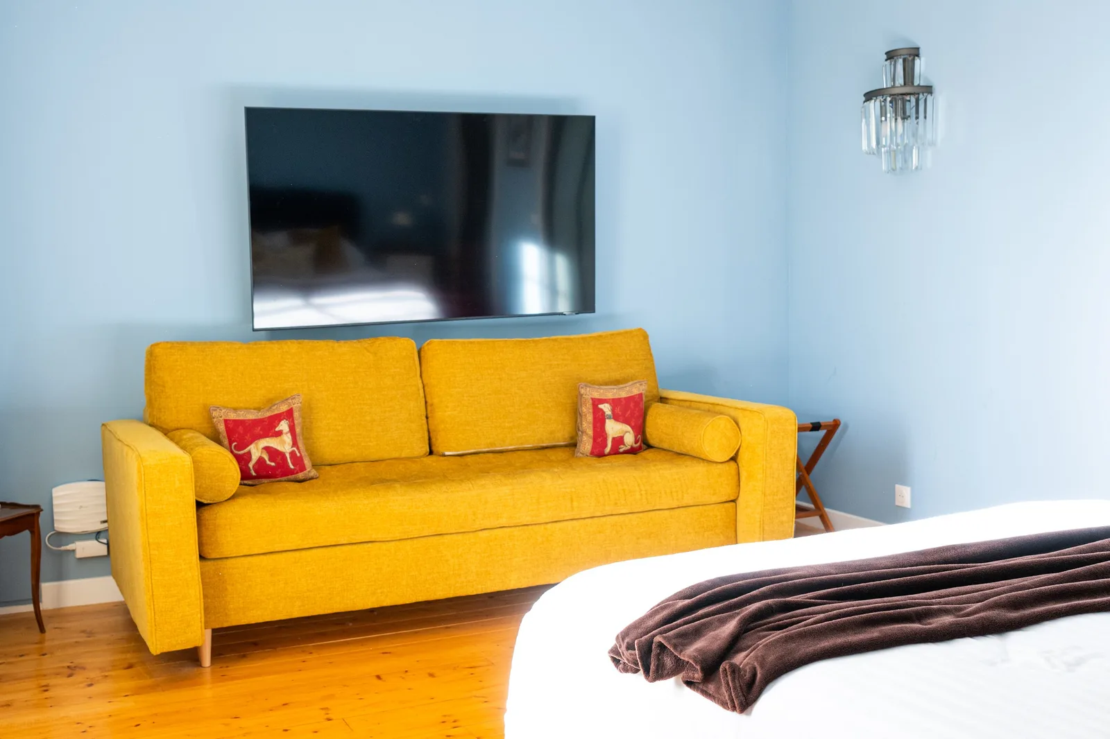 Coin salon de la Suite La Comtesse avec canapé-lit jaune moutarde, coussins brodés à motif de lévrier, télévision murale et mur bleu ciel.