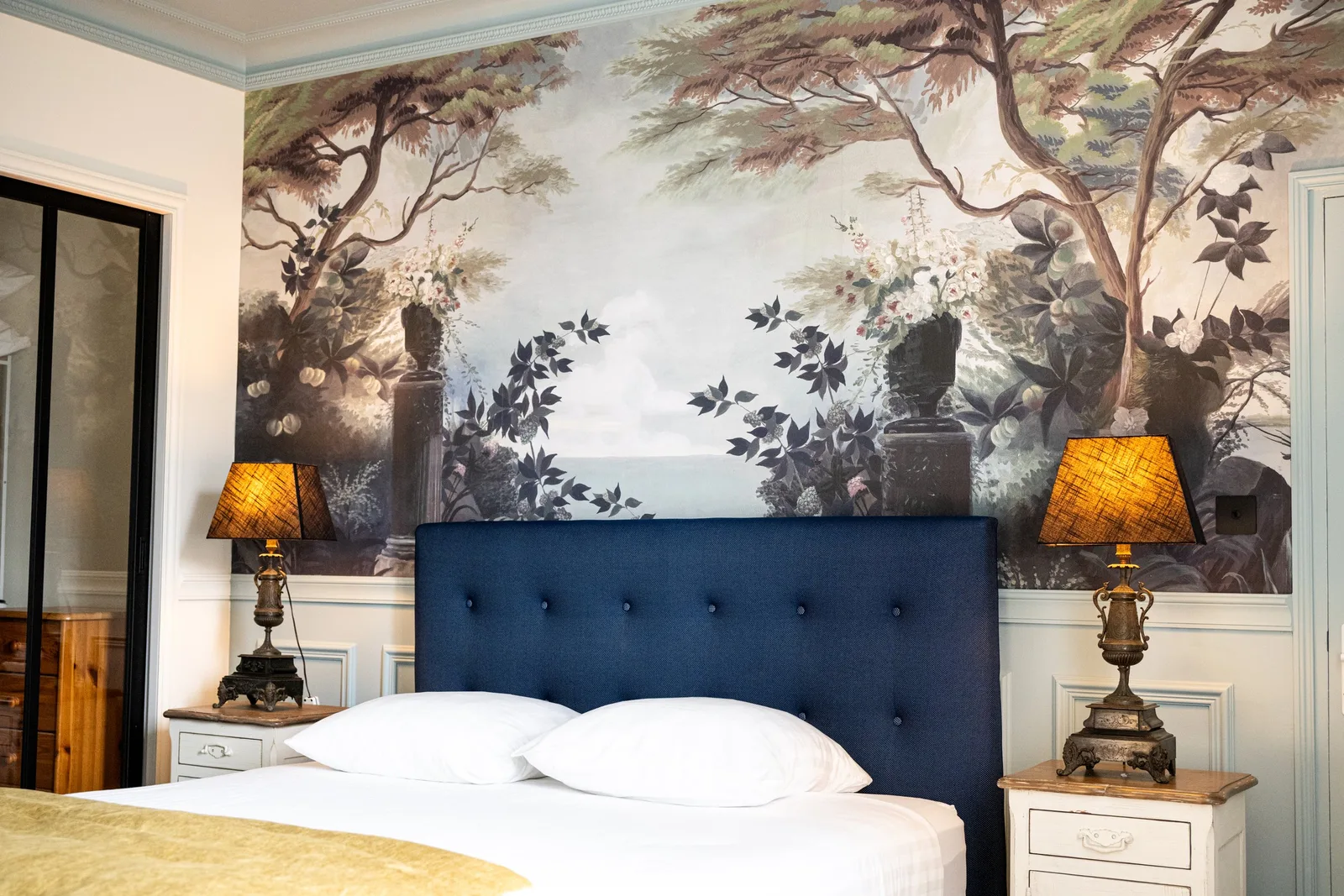 Tête de lit capitonnée bleu marine de la Suite La Comtesse encadrée de deux tables de chevet anciennes et de lampes à abat-jour tressé, devant le papier peint panoramique arboré.