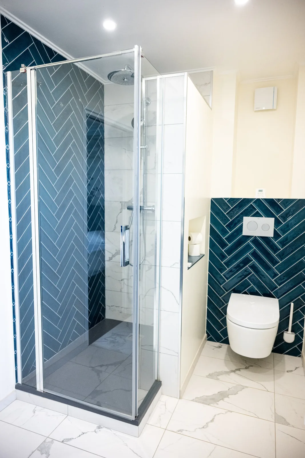 Douche à l'italienne carrelée en faïence bleu canard à motif chevron, avec douche de pluie, dans la salle de bain de la Suite La Comtesse ; WC japonais suspendu visible sur la droite.