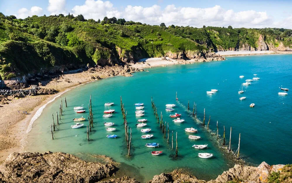 Vue aérienne du petit port de Gwin Zegal en Bretagne, bateaux amarrés à des pieux de bois plantés dans le sable au pied de hautes falaises verdoyantes.