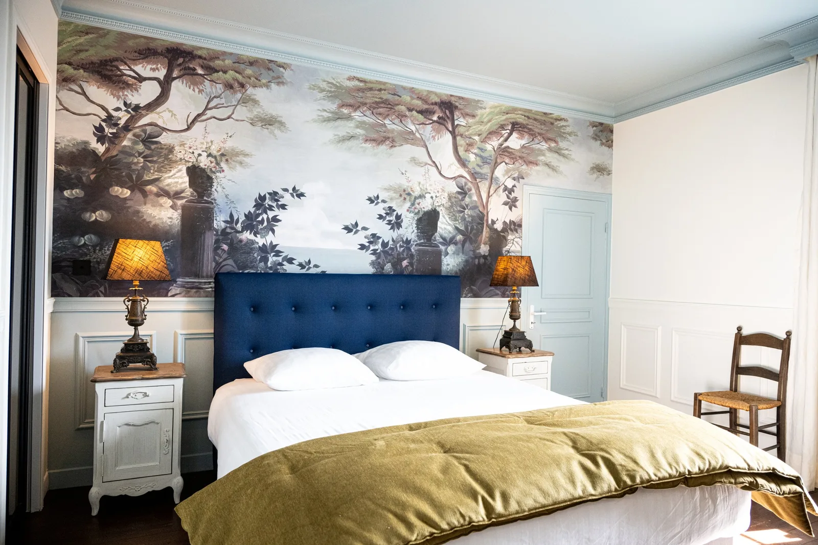 Vue d'ensemble de la Suite La Comtesse : lit double à tête de lit bleu marine capitonnée devant un papier peint panoramique représentant un jardin arboré, encadré de deux tables de chevet anciennes et de leurs lampes.