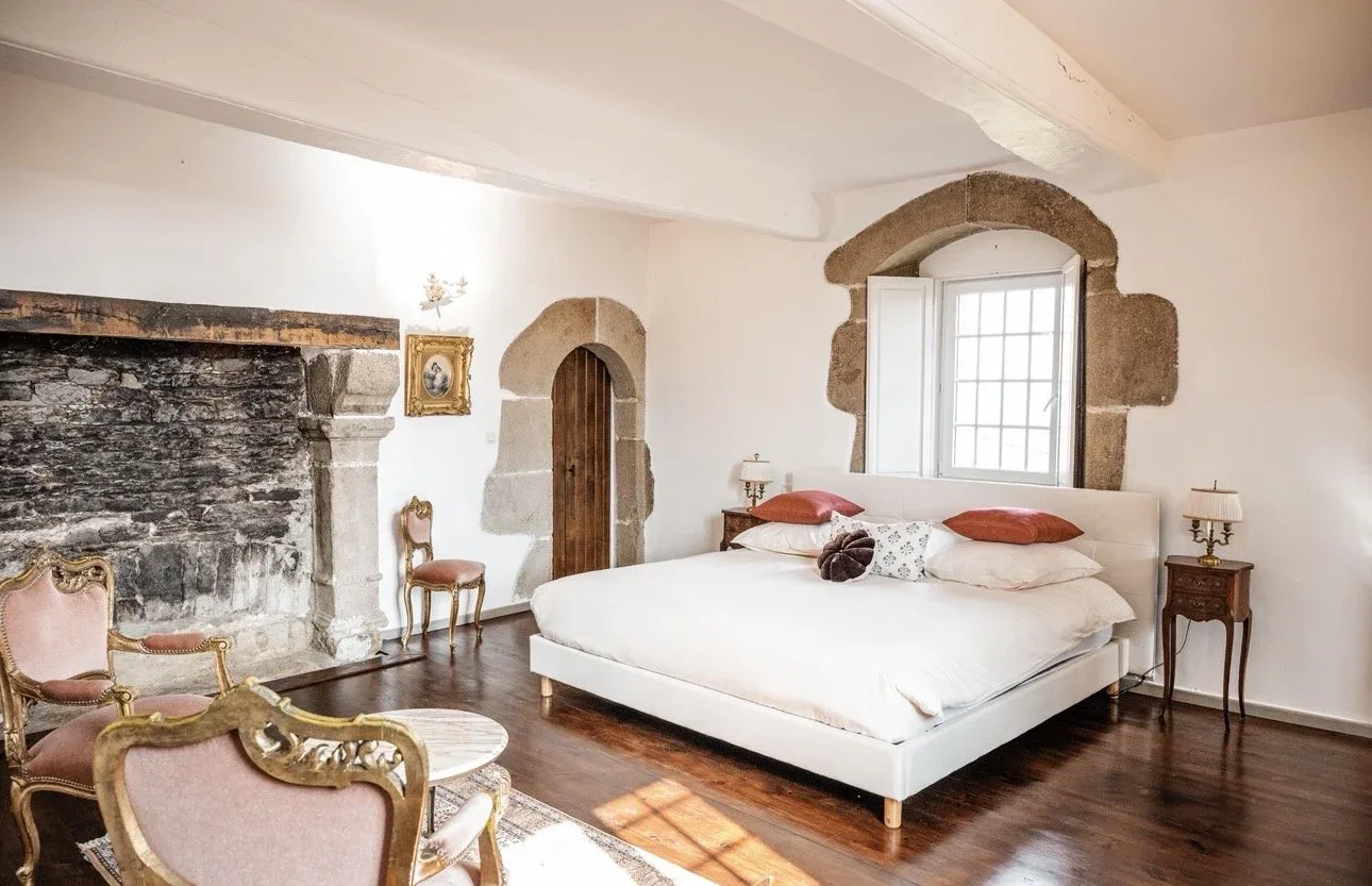 Suite du Maître vue à travers une arche en pierre de granit, avec fenêtre à petits carreaux encadrée de pierre, lit blanc capitonné et fauteuil ancien doré.