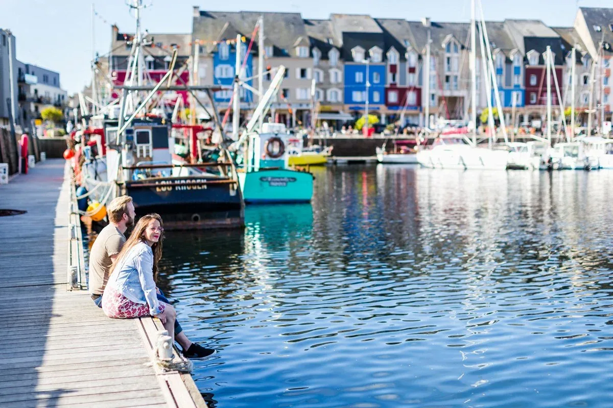 Le port de Paimpol en Bretagne : bateaux de pêche et voiliers amarrés devant les maisons colorées du quai, un couple assis sur le ponton.