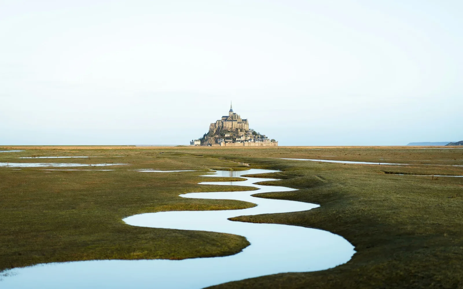Vue du Mont Saint-Michel émergeant de la baie, avec un chenal serpentant à travers les prés salés.