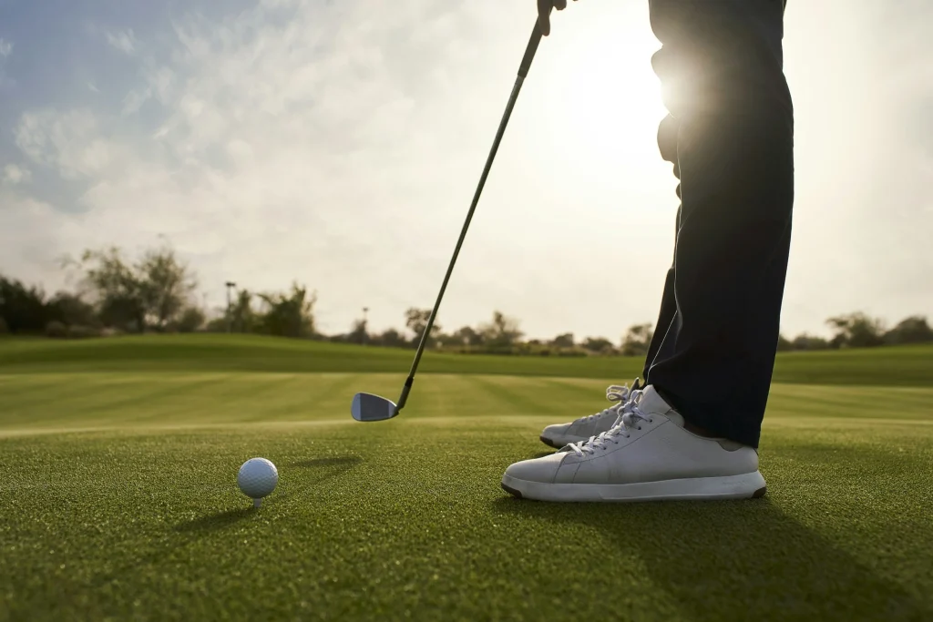 Golfeur prêt à frapper une balle posée sur un tee, gros plan sur ses chaussures et son club sur un green baigné de soleil