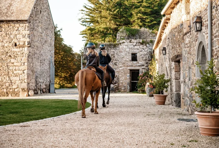 Deux cavalières à cheval de dos, quittant au pas la cour pavée du Manoir bordée de bâtiments en pierre, en lumière dorée de fin de journée