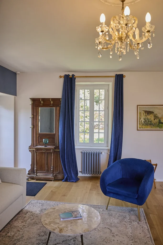 Salon avec une cheminée en bois, une fenêtre avec des rideaux bleus, un fauteuil bleu, un canapé beige, et un tapis avec une table en marbre. Un lustre en cristal au plafond.