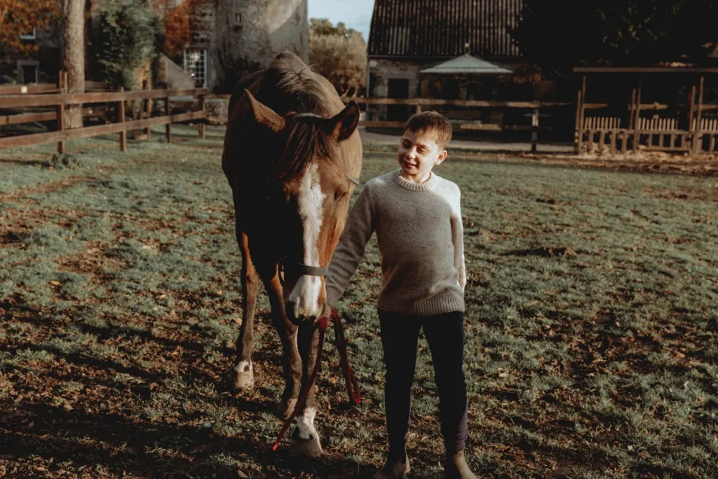 Un garçon jeune en pull gris marche avec un cheval dans un pré ensoleillé, entouré de clôtures en bois et de bâtiments en arrière-plan.