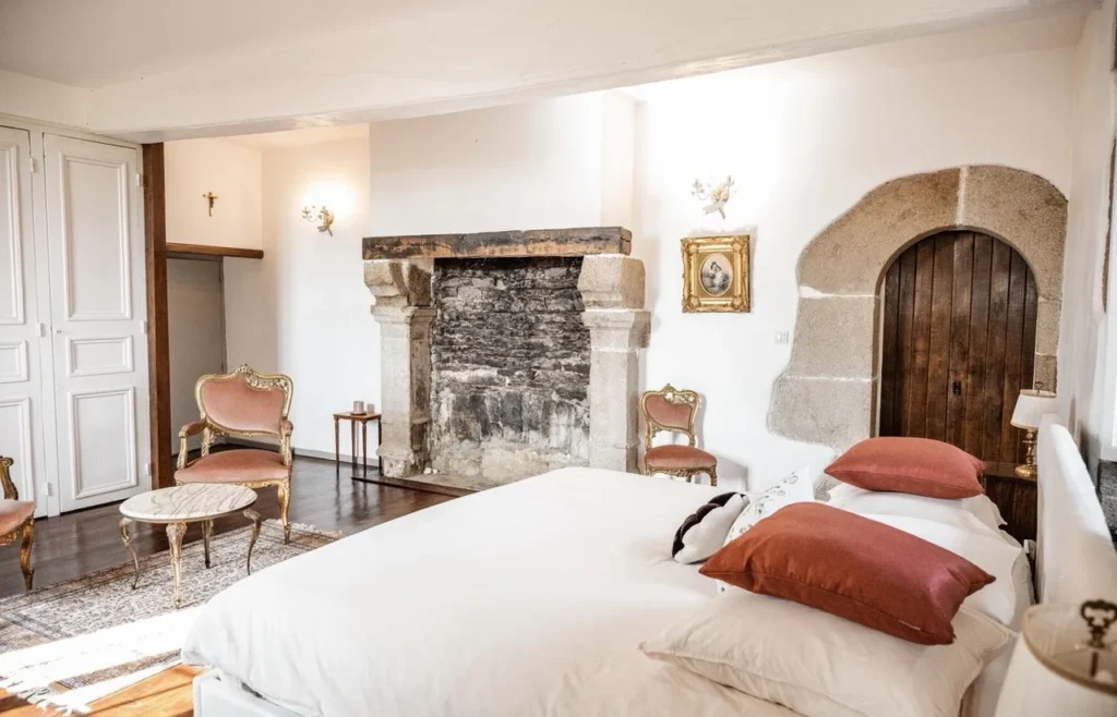 Chambre élégante avec cheminée en pierre, lit avec oreillers rouges, fauteuils en velours rose, petite table ronde en marbre, rotin.