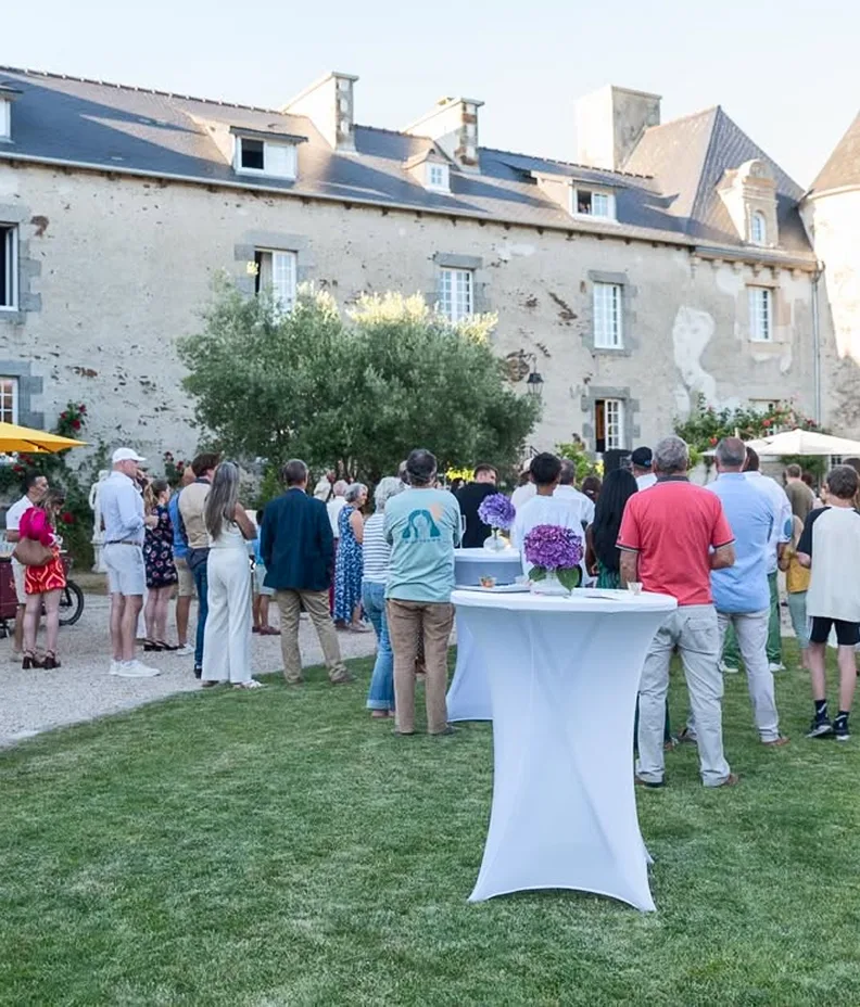 Un rassemblement de personnes lors d'un événement en plein air devant une vieille bâtisse en pierre, avec des tables décorées de fleurs violettes.
