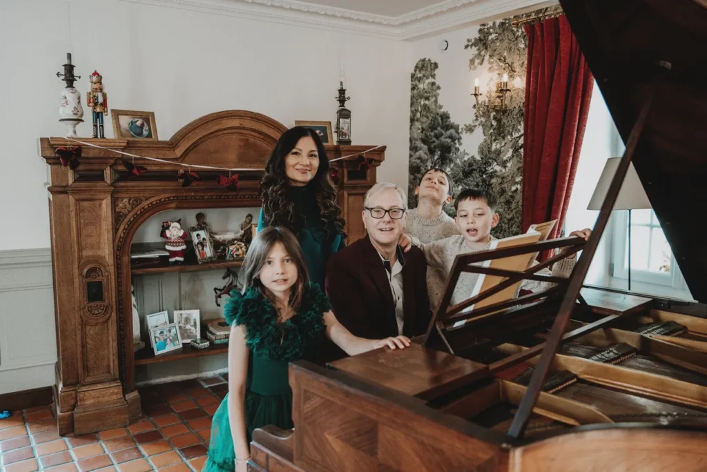 Une famille avec sept membres, comprenant deux adultes et cinq enfants, pose autour d'un piano à queue en bois dans un salon décoré pour Noël, avec des photos de famille, des décorations et des rideaux rouges.