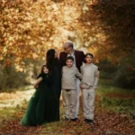 Une famille de cinq personnes marche ensemble dans un bois en automne, entourée d'arbres aux feuilles orangées, souriant et profitant de l'instant.