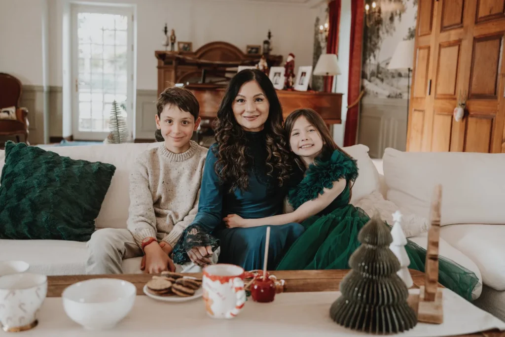 Une femme et deux enfants assis sur un canapé dans un salon décoré pour Noël, avec des biscuits et des décorations de Noël sur la table devant eux.