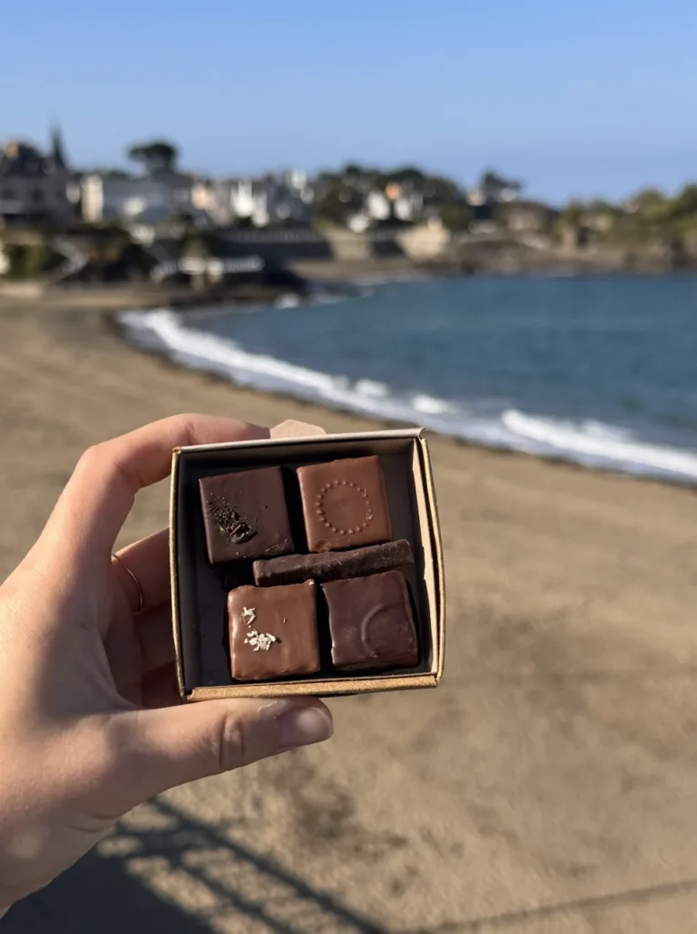 Une main tient une boite de chocolats en face d'une plage et de la mer avec des bâtiments en arrière-plan.