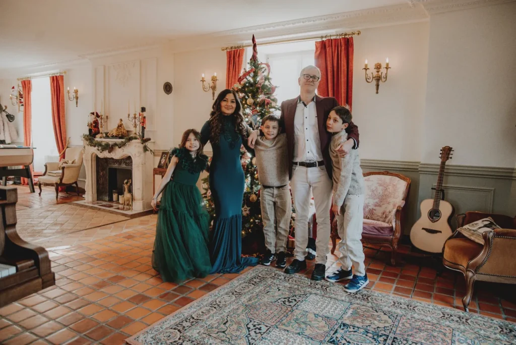 Une famille posant devant un sapin de Noël décoré dans un salon élégant. Il y a deux adultes et trois enfants, tous souriants. La pièce est décorée avec des guirlandes, une cheminée, des rideaux rouges et une guitare posée contre un fauteuil.