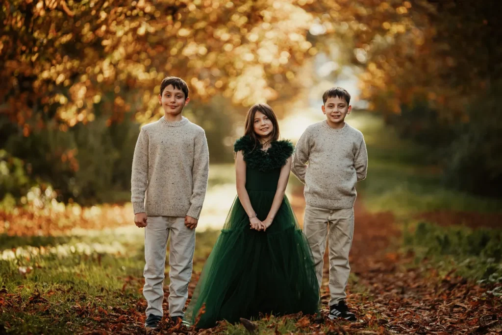 Trois enfants posant dans un parc en automne, deux garçons en pull beige et une fille en robe verte avec des feuilles mortes au sol et des arbres aux feuilles orangées en arrière-plan.